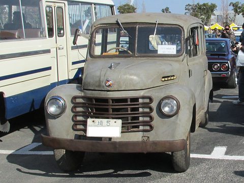 Weathered grey light van seen from the front with a horizontal grille and round headlamps, a bus beside it. Opens a larger view