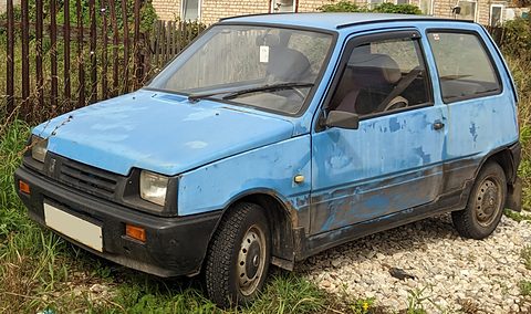 Faded pale blue small hatchback with rust and mud on its sills, parked on gravel by a wooden fence and tall weeds. Opens a larger view