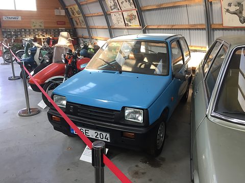 Blue small hatchback behind a red rope barrier in a museum shed, motorcycles lined up along the wall. Opens a larger view