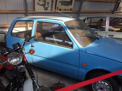 Blue small hatchback in a museum shed, a red motorcycle in front of it and a white saloon behind. Opens a larger view