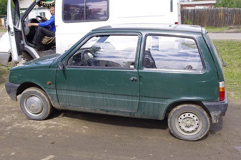 Side view of a muddy dark green small hatchback on a dirt yard, a white van with its door open behind. Opens a larger view