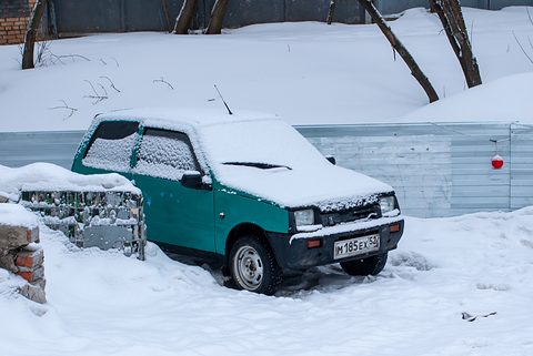 Small green hatchback with snow on its roof and bonnet, parked in deep snow beside a corrugated fence. Opens a larger view