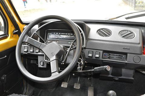 Black plastic dashboard and three-spoke wheel of a small car, a frame of hand levers mounted under the column.