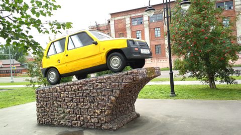 A yellow Oka mounted at an angle on a stone-filled plinth on a lawn, a red-brick factory building behind.