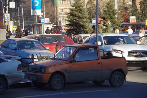 Orange Oka with its rear roof cut away into a small open bed and a roll bar, parked among cars in a city street. Opens a larger view