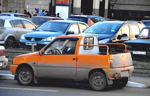 Side view of an orange and grey Oka with a cut-down open rear and a tubular roll bar, other cars parked behind. Opens a larger view