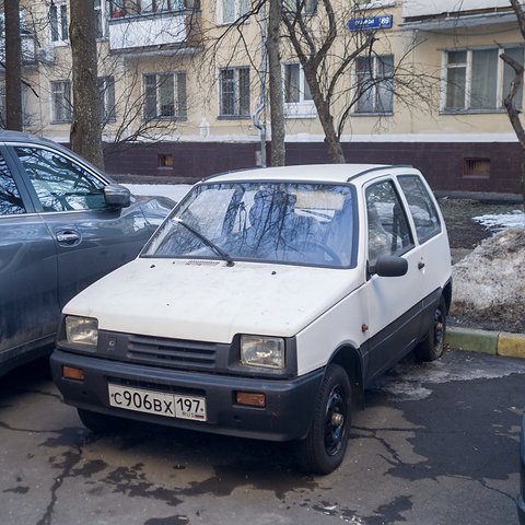 White small hatchback with a dark lower body parked by a snowbank in front of a pale apartment block. Opens a larger view