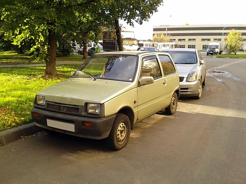 Beige small hatchback seen almost head-on, parked on a leafy street with a silver car behind it. Opens a larger view
