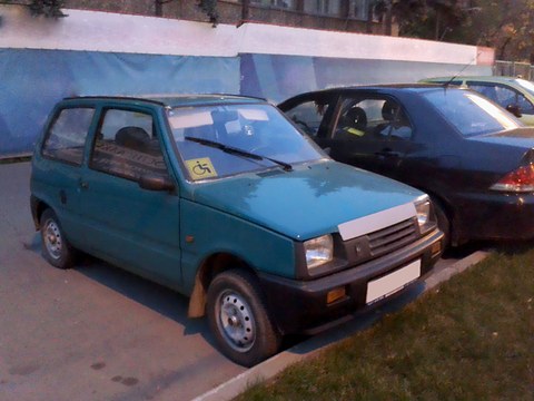 Dark green small hatchback with a white patch over its grille and a disabled-driver sticker, parked by a blue fence. Opens a larger view