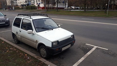 White small hatchback with a black roof rack parked in a marked bay beside a wide road. Opens a larger view