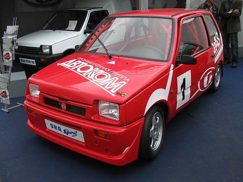 Red small hatchback with a roll cage, sponsor lettering and a number 1 on the door, on a show stand. Opens a larger view