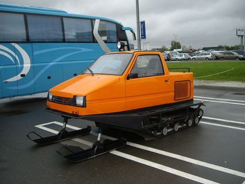 Orange snowmobile with a car-like cab, rubber tracks and front skis, parked in a car park beside a blue coach. Opens a larger view