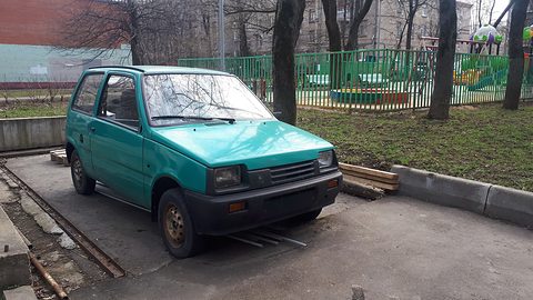Turquoise small three-door hatchback on a concrete pad beside timber and grass, a green fence and playground behind. Opens a larger view