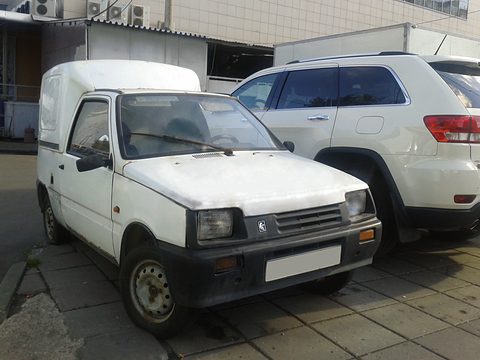 White Oka with a high-roofed van body, black bumper and plain grille, parked on a pavement beside a large SUV. Opens a larger view