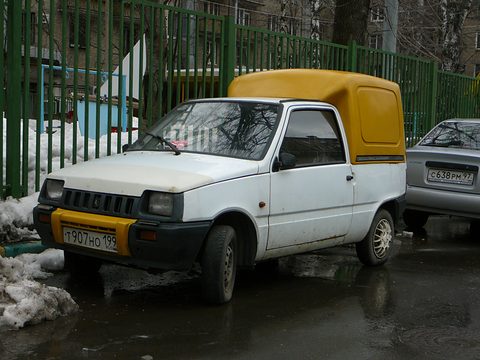 White Oka with a tall yellow plastic box over its rear, parked by a green fence with melting snow around it. Opens a larger view