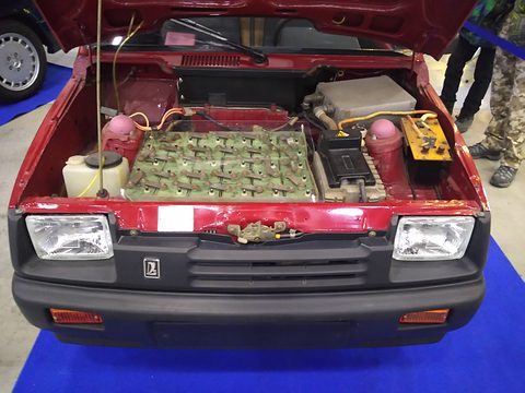 Open engine bay of a red car packed with a grid of battery cells and wiring, round headlamps either side. Opens a larger view