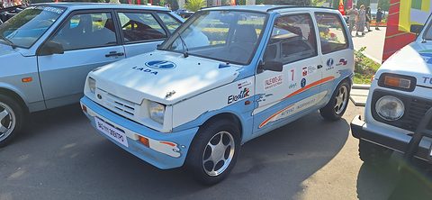 White three-door Oka with pale blue splash graphics, Electric lettering and rally stickers, parked among other Ladas outdoors. Opens a larger view