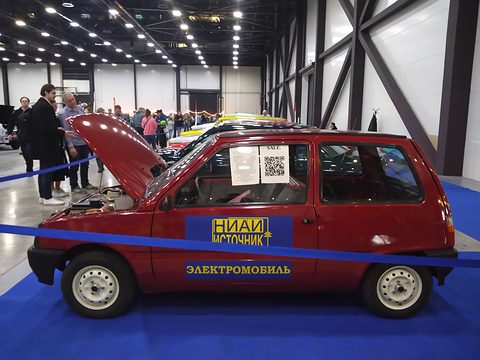 Side view of a red small hatchback with its bonnet raised, ELECTROMOBILE lettering on the door, behind a blue rope. Opens a larger view