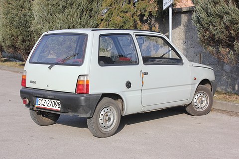 Pale grey small three-door hatchback from the rear left, parked on a village road under a wooded bank. Opens a larger view
