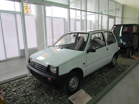 White small hatchback on a bed of pebbles in a glass-walled museum hall, a dark off-roader behind. Opens a larger view