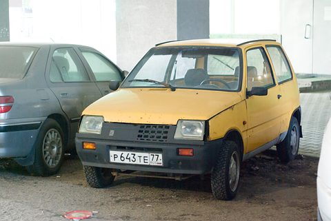 Dusty yellow small hatchback with black bumpers parked under a building beside a grey saloon. Opens a larger view