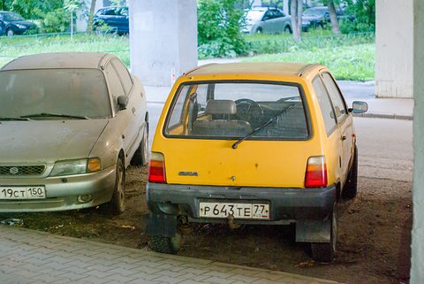 Rear of a yellow small hatchback with a black bumper and red tail lamps, parked beside a dusty grey saloon. Opens a larger view