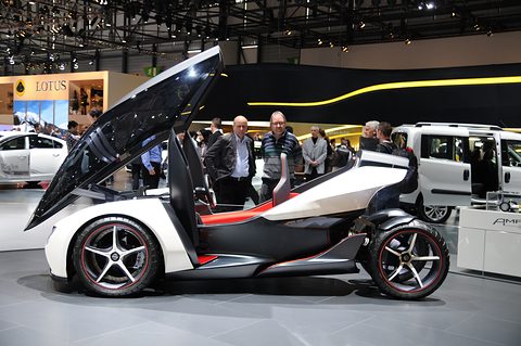 Side view of a white and black tandem concept with its canopy lifted high, visitors and stands behind. Opens a larger view