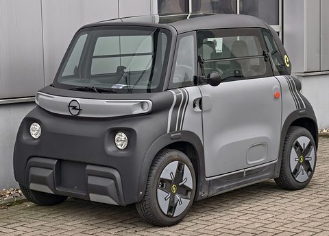Grey and black cube-shaped two-seater with an Opel badge and striped decals, parked on herringbone paving by a metal building. Opens a larger view