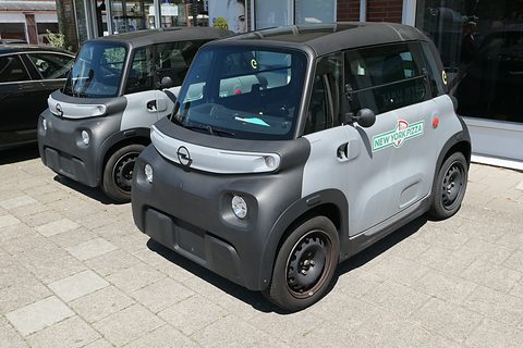 Two grey and black cube-shaped cars parked on paving outside a shop window, one with New York Pizza lettering. Opens a larger view