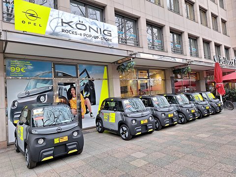 Six grey and yellow cube-shaped cars lined up on a pavement in front of a shop with an Opel König sign.