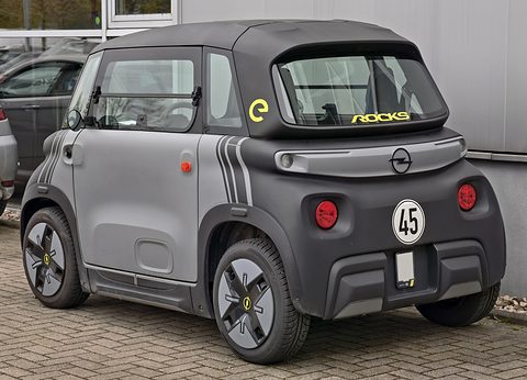 Rear of a grey and black cube-shaped two-seater with a round 45 sticker and Rocks lettering on its rear window. Opens a larger view