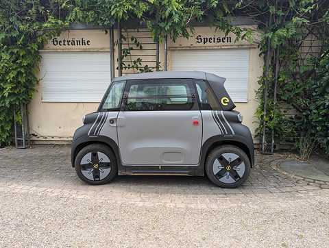 Grey cube-shaped two-seater with black stripes seen side-on on gravel in front of a closed kiosk with shuttered windows. Opens a larger view