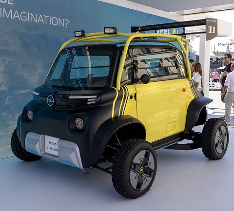 Yellow and black raised cube-shaped car with roof lamps, a roll cage and off-road tyres in front of a sky-blue screen. Opens a larger view