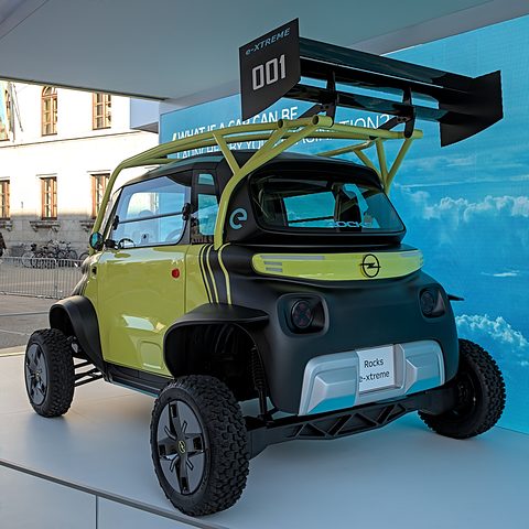 Rear of a yellow and black raised two-seater with a tall rear wing, a roll cage and off-road tyres, under an outdoor canopy. Opens a larger view