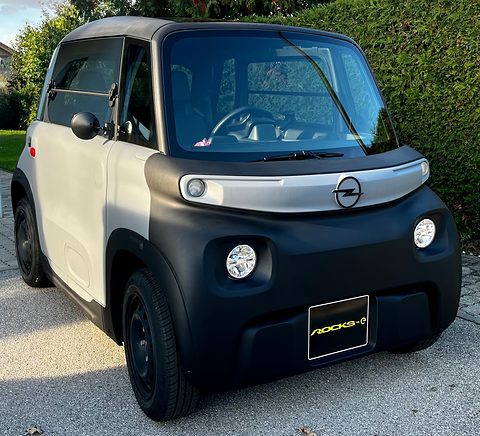 White and black cube-shaped two-seater with an Opel badge and a Rocks-e plate, parked on gravel by a hedge. Opens a larger view