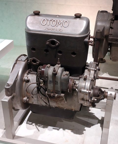 A grey cast engine on a stand, the word OTOMO cast into its top cover, with a magneto and flywheel housing.