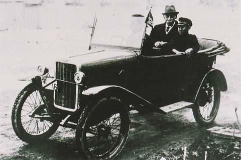 A small open tourer with wire wheels and a flat radiator, a pennant on the windscreen, two men seated in its rear. Opens a larger view