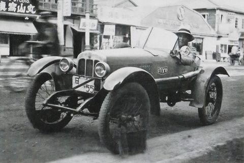 A small open car with a flat radiator and wire wheels on a shop-lined street, a driver in a hat aboard. Opens a larger view