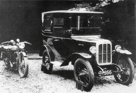 A tall black closed car with a white-framed radiator and wire wheels on gravel, a motorcycle parked beside it.
