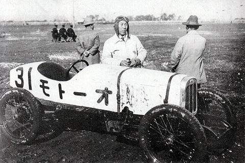 A white single-seat racing car with a pointed tail and spoked wheels, the name in katakana on its flank, men standing behind.