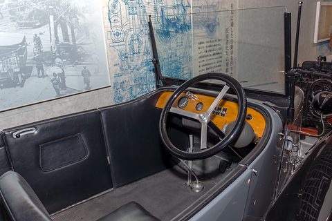 A black three-spoke steering wheel before a varnished wooden dashboard with small gauges and a windscreen frame. Opens a larger view