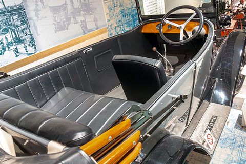 Pleated black leather front and rear seats, a steering wheel and a varnished dashboard inside a grey open car. Opens a larger view