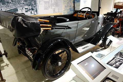Rear three-quarter view of a grey tourer with its black hood folded over the tail and a spare wheel behind. Opens a larger view