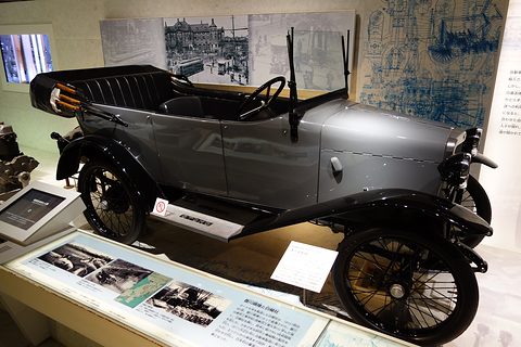 Side view of a grey open tourer with four seats, cycle-type wings and black wire wheels, period street photographs behind. Opens a larger view