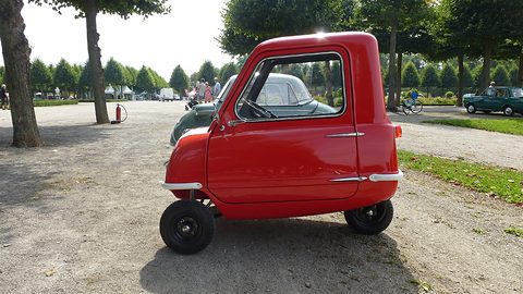 Tiny red car in profile on a gravel avenue lined with trees. Opens a larger view