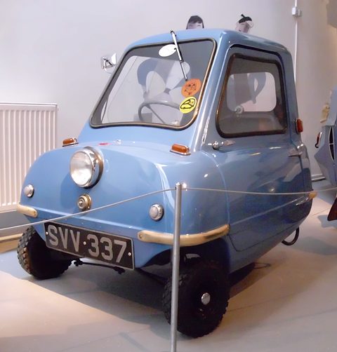 Tiny pale blue car seen from the front behind a thin rope barrier in a white museum. Opens a larger view