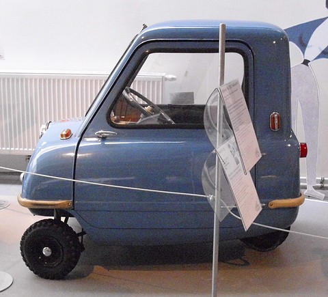 Tiny blue car in side view with its one door, a placard on a stand in front