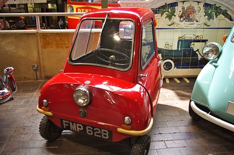 Tiny red car seen from the front on a tiled floor, a tiled mural of a vintage car behind. Opens a larger view