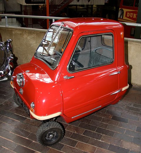 Tiny red single-seat car on a wooden floor, seen from the front left with the nearside door. Opens a larger view