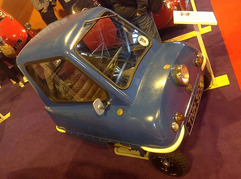Tiny blue car seen from above on a red carpet, a yellow display stand behind. Opens a larger view